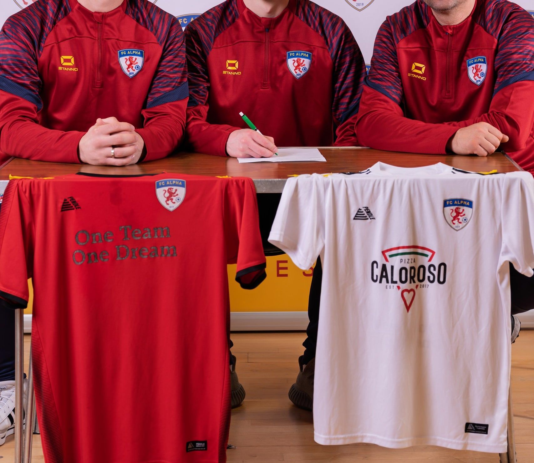 FC Alpha club officials and a player at the signing table with the new Caloroso Pizza shirts